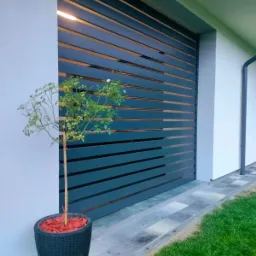 Nowoczesna, pozioma brama wjazdowa z metalowych profili, oświetlona od wewnątrz, z małym drzewkiem w donicy z czerwonymi kamykami po lewej stronie, na tle jasnej elewacji domu i zielonego trawnika.