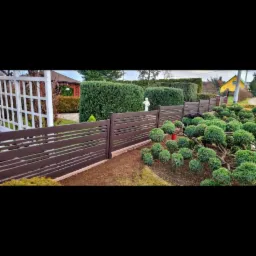 Nowoczesne, brązowe ogrodzenie z poziomymi szczelinami, oddzielające zadbany ogród z kulistymi krzewami od trawnika, w tle biała altana i rzeźba.