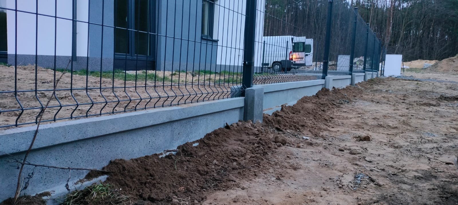 Częściowo ukończone ogrodzenie z paneli z podmurówką betonową, widoczny samochód dostawczy w tle, teren po budowie.