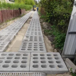 Układanie ażurowych płyt betonowych na drodze dojazdowej, z widocznym minikoparką w tle. Prace ziemne w toku, teren przygotowany pod dalsze utwardzanie.