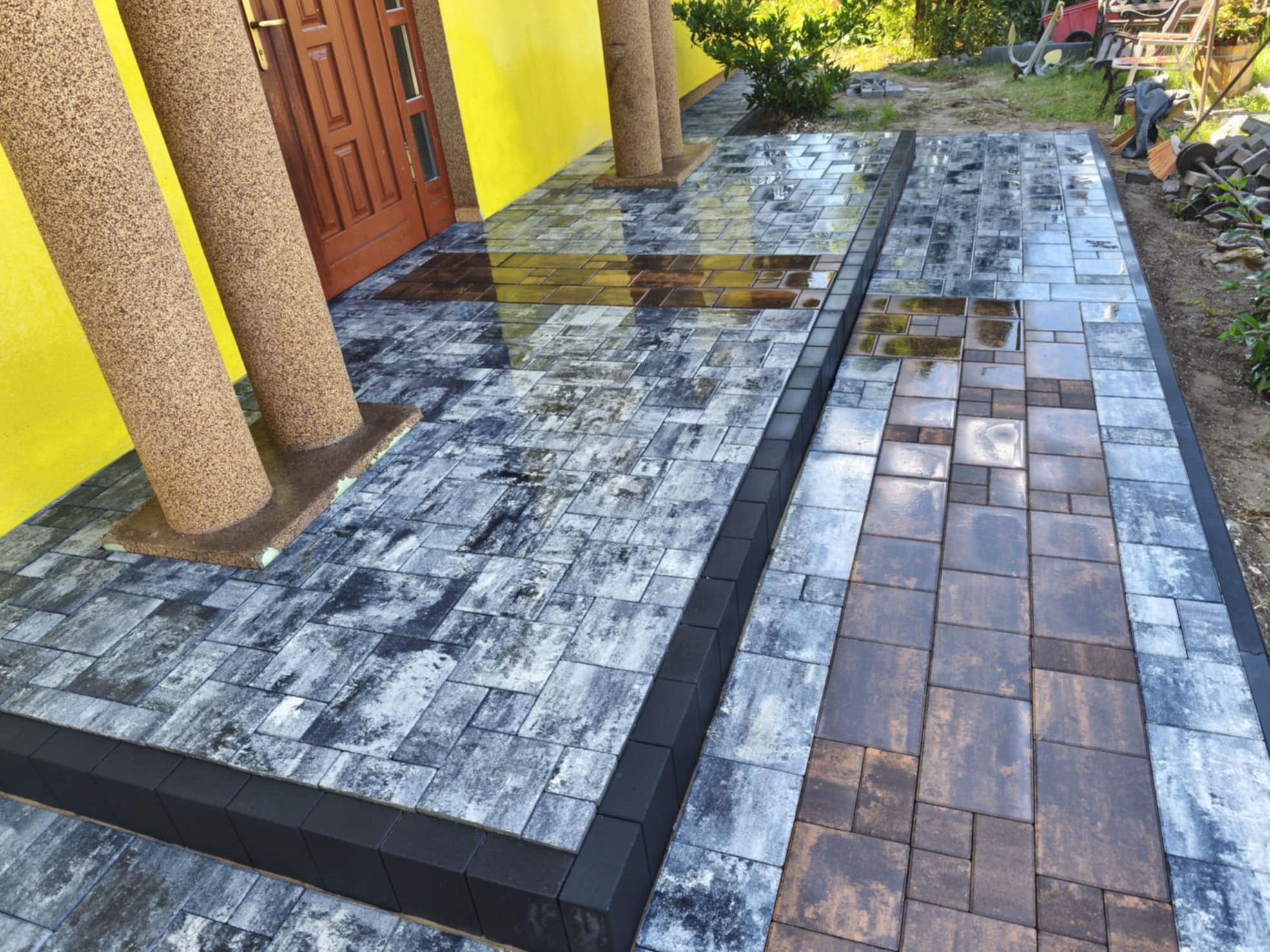 Ułożona kostka brukowa w dwóch kolorach, szara i brązowa, wykończona czarnym obrzeżem, prowadząca do drzwi budynku. Widoczne kolumny przy wejściu.