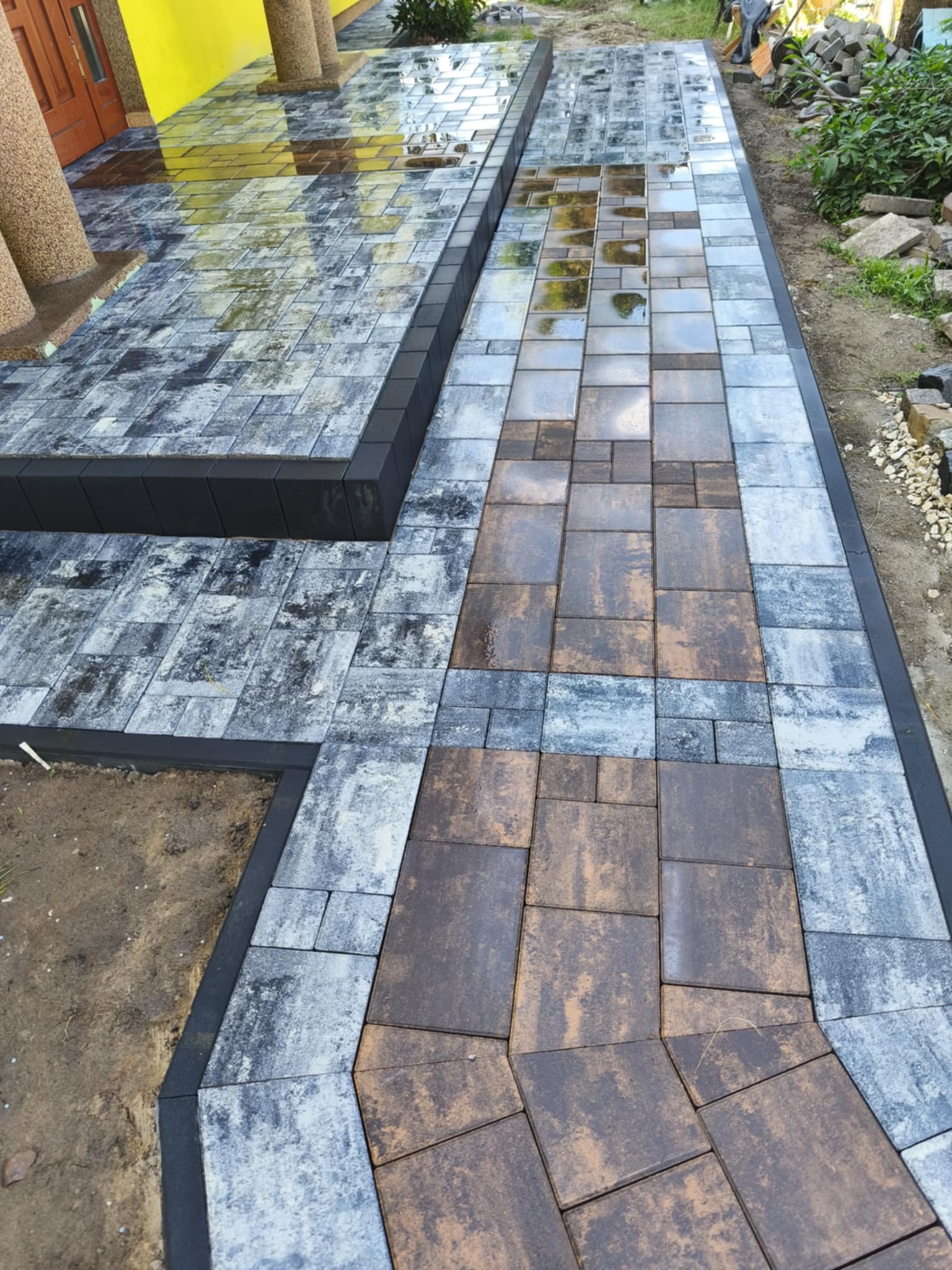 Świeżo ułożona, mokra ścieżka z kostki brukowej w odcieniach szarości i brązu, z czarnym obrzeżem, prowadząca do kolumnady domu. Widoczne odbicia światła w kałużach.