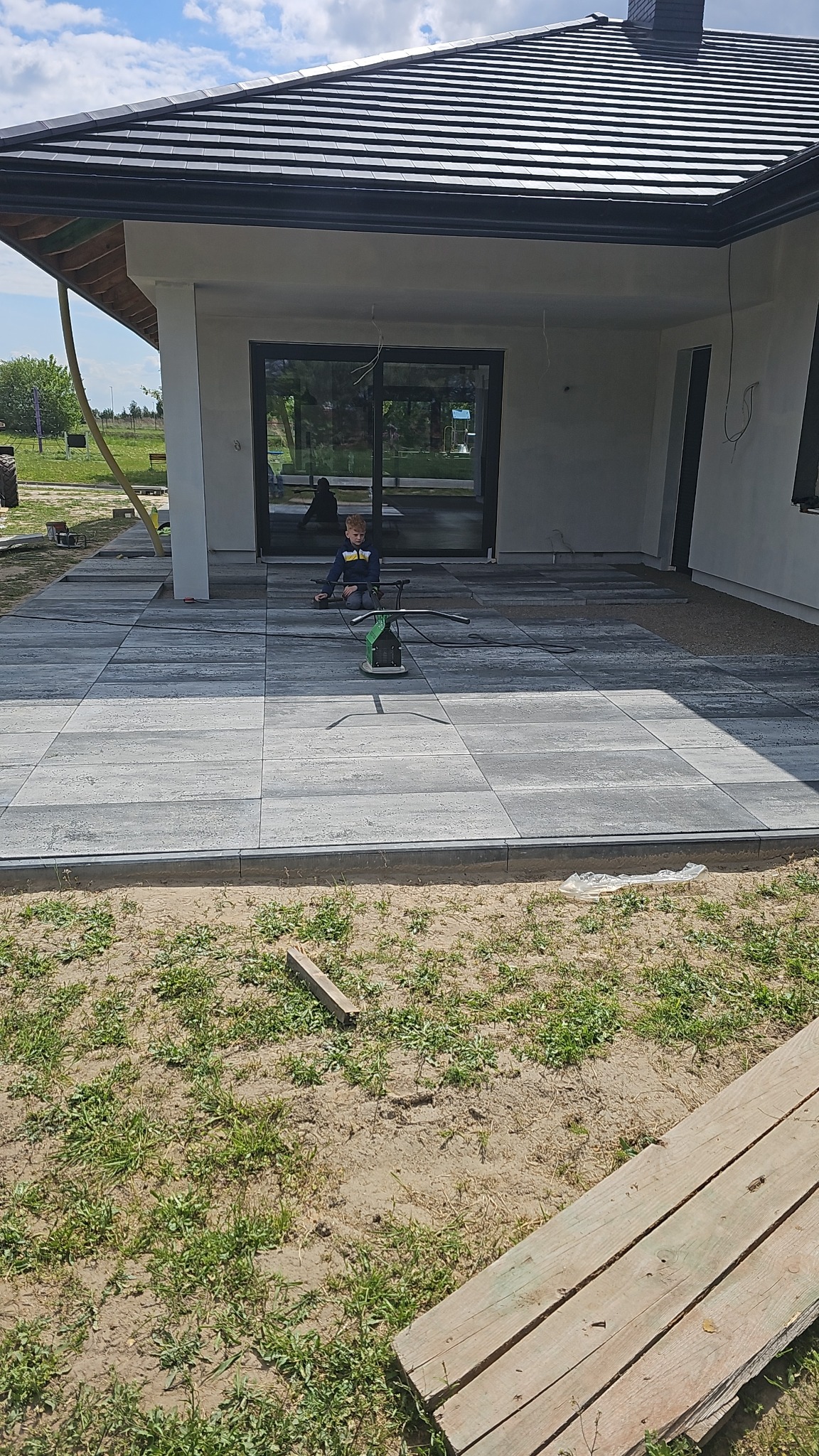 Ułożona szara kostka brukowa na tarasie domu jednorodzinnego. Widoczna zagęszczarka, chłopiec siedzący na kostce i trawnik w tle. Nowoczesna architektura.