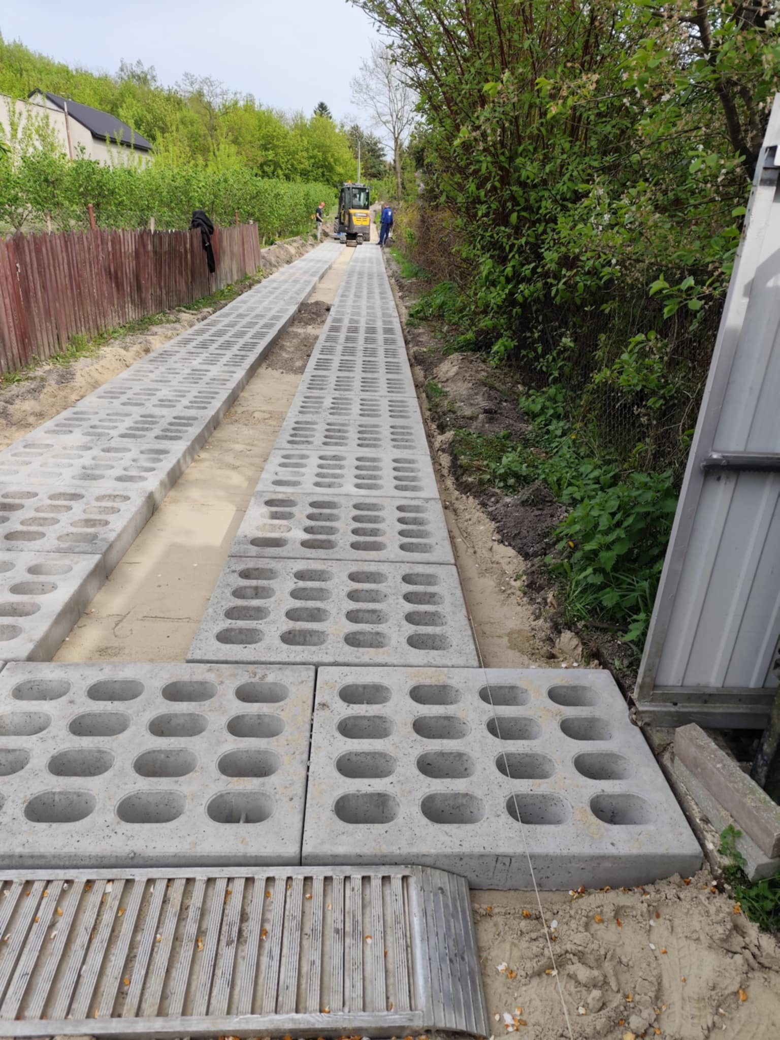 Układanie ażurowych płyt betonowych na drodze dojazdowej, z widocznym minikoparką w tle. Prace ziemne w toku, teren przygotowany pod dalsze utwardzanie.