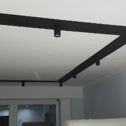 Czarny, geometryczny sufit podwieszany z punktowym oświetleniem LED w nowoczesnym wnętrzu, widoczne okno z folią ochronną i częściowo zakryte meble.