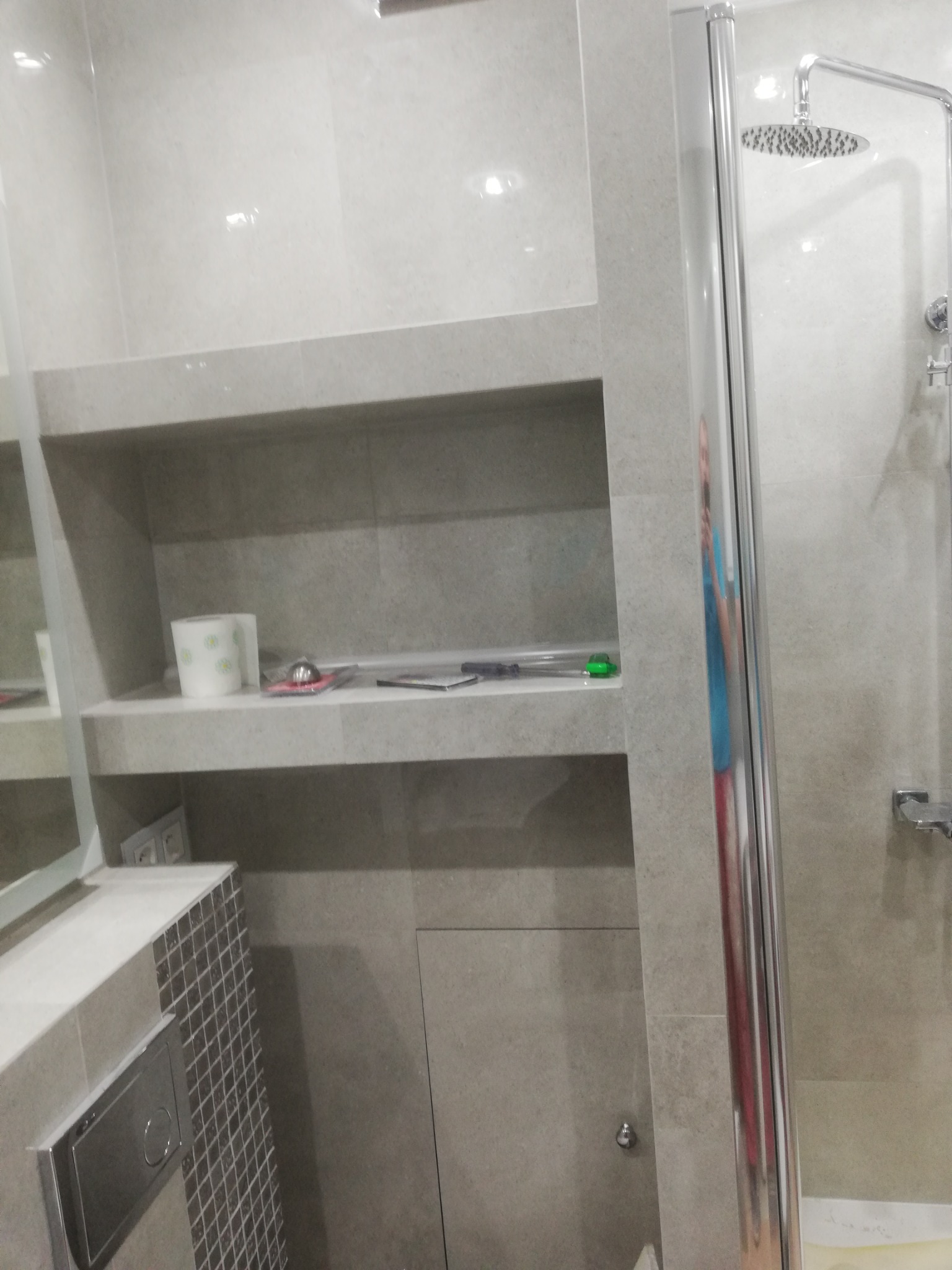 Wykończona łazienka w odcieniach szarości, z kabiną prysznicową typu walk-in, półkami wnękowymi z akcesoriami i mozaiką przy toalecie podtynkowej.