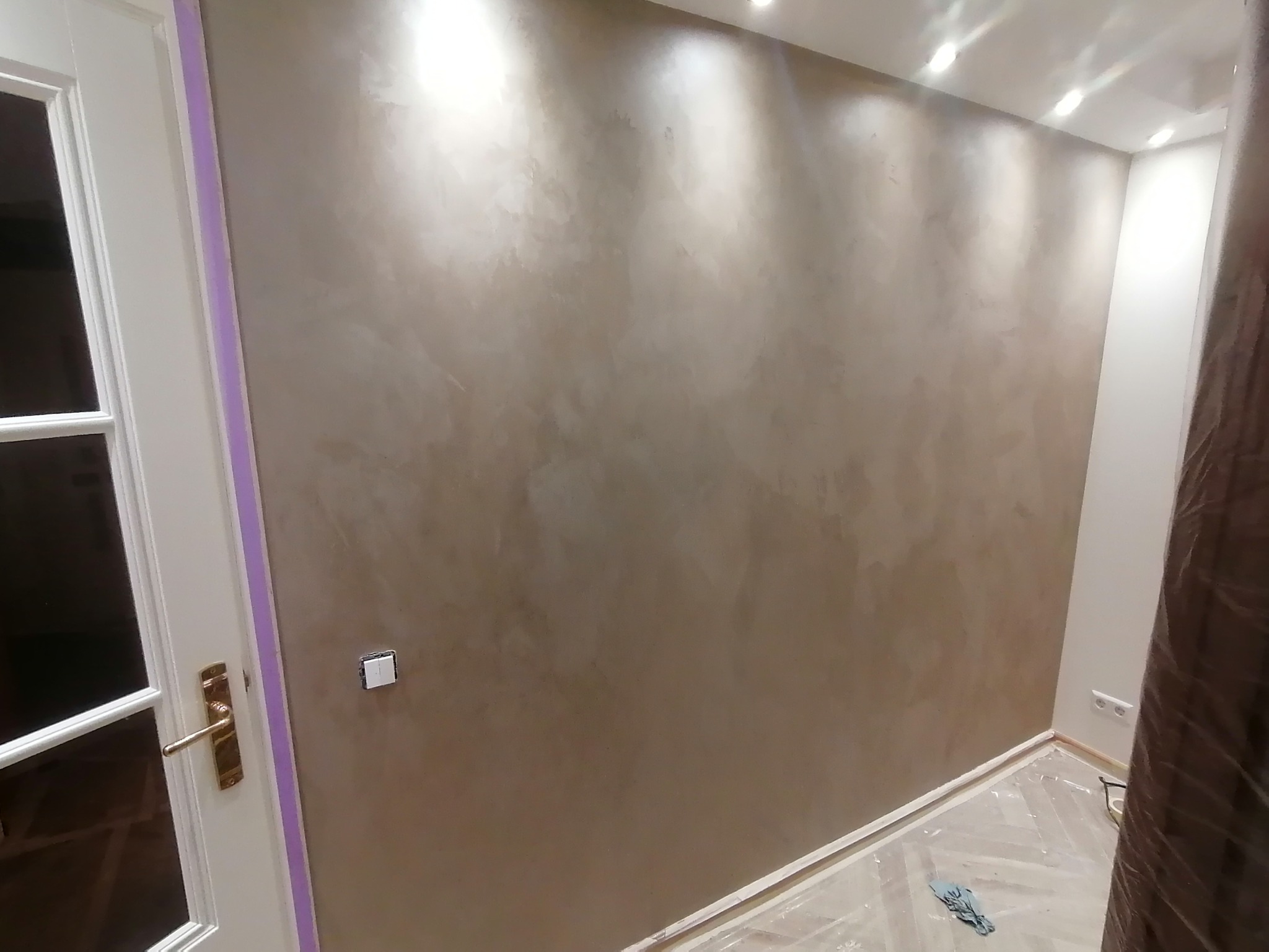 Ściana w pokoju po nałożeniu dekoracyjnej, beżowej powłoki z efektem cieniowania, widoczne zabezpieczone folią podłoga i listwy przypodłogowe, oraz fragment drzwi z taśmą malarską.