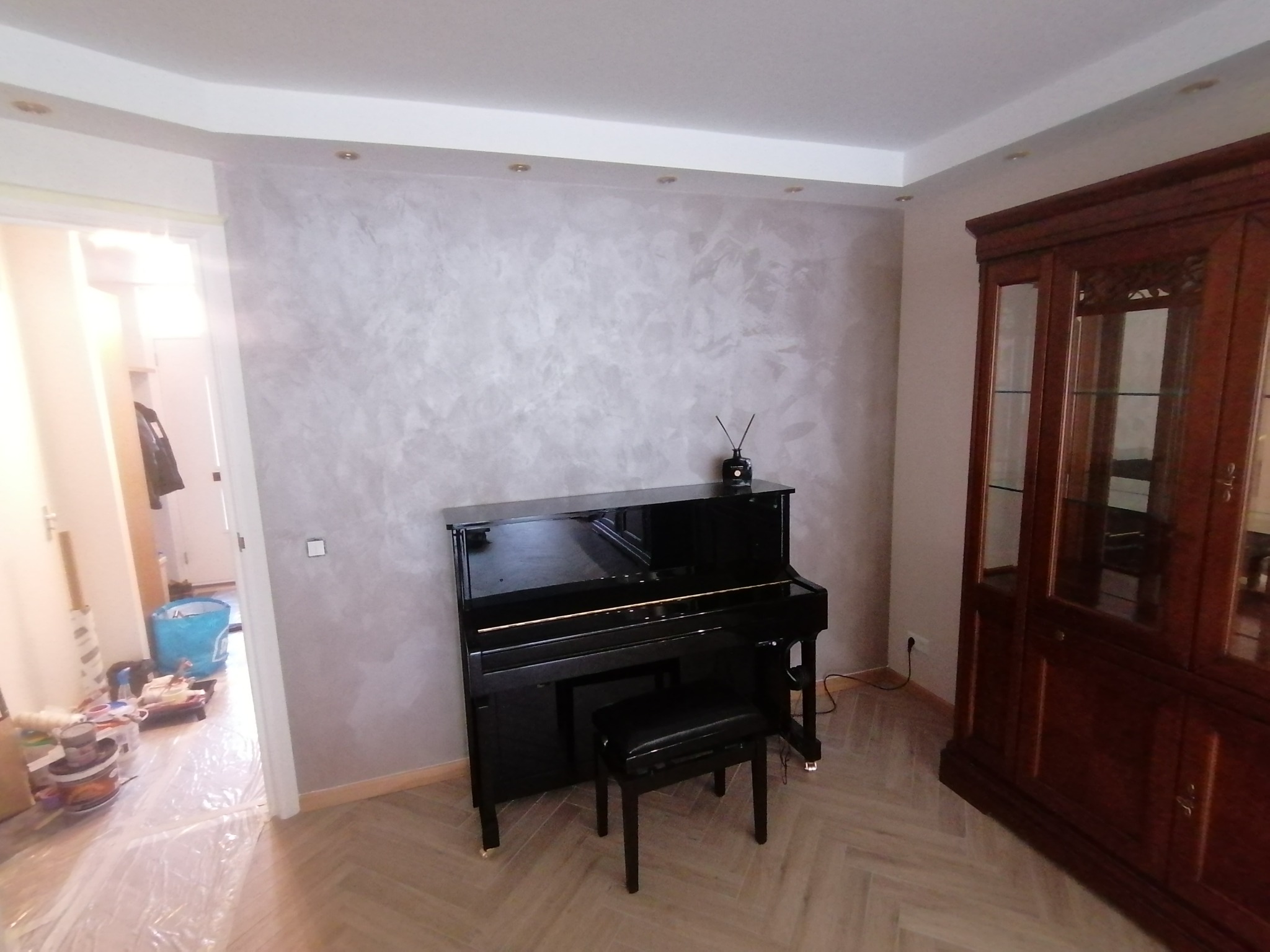 Ściana z dekoracyjną szarą fakturą w salonie z czarnym pianinem i drewnianą witryną, widoczne przygotowania do prac wykończeniowych w przejściu.