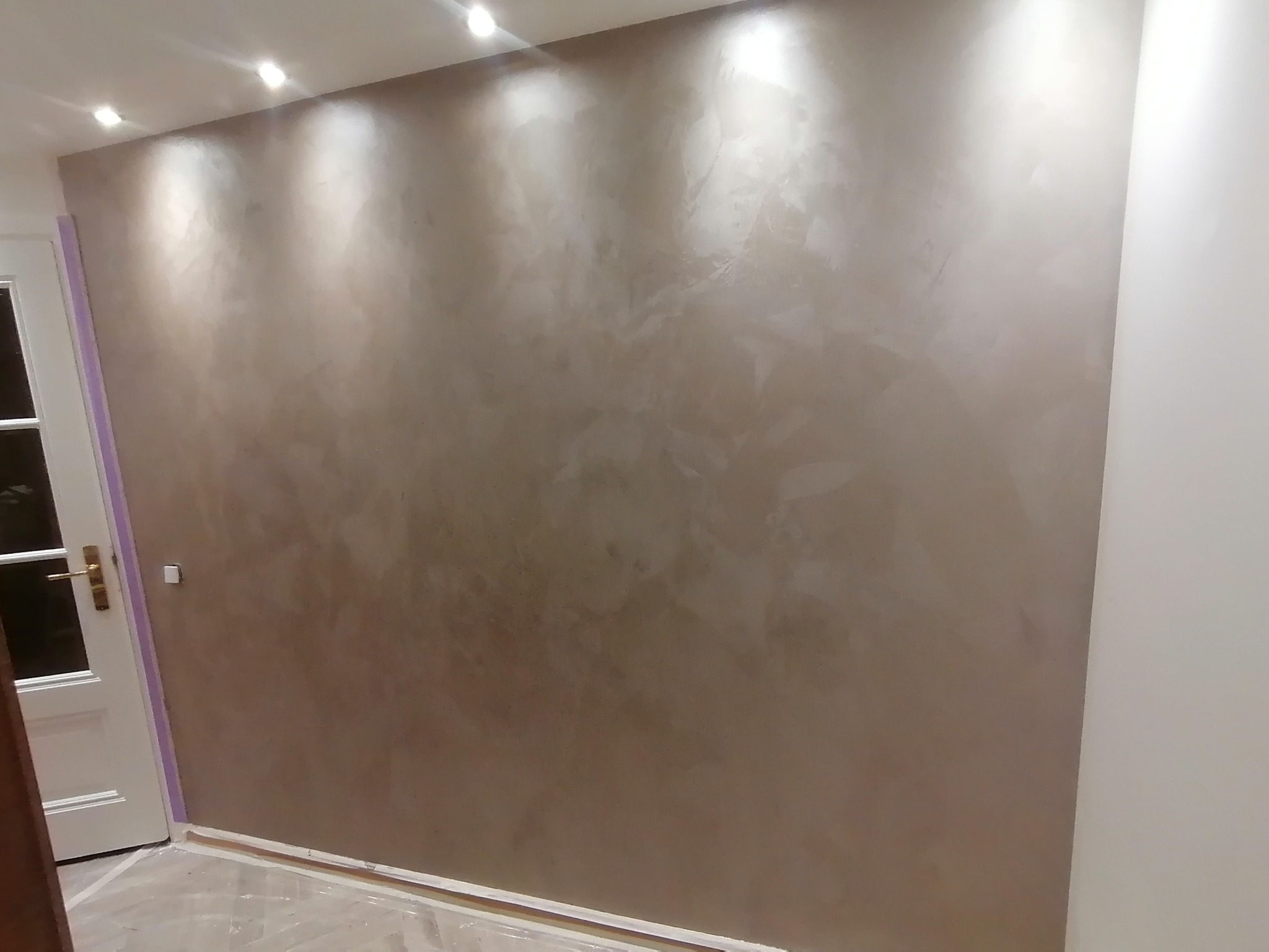 Ściana z dekoracyjną, beżową fakturą, oświetlona punktowymi lampami sufitowymi, zabezpieczona folią podłoga i listwa przypodłogowa.