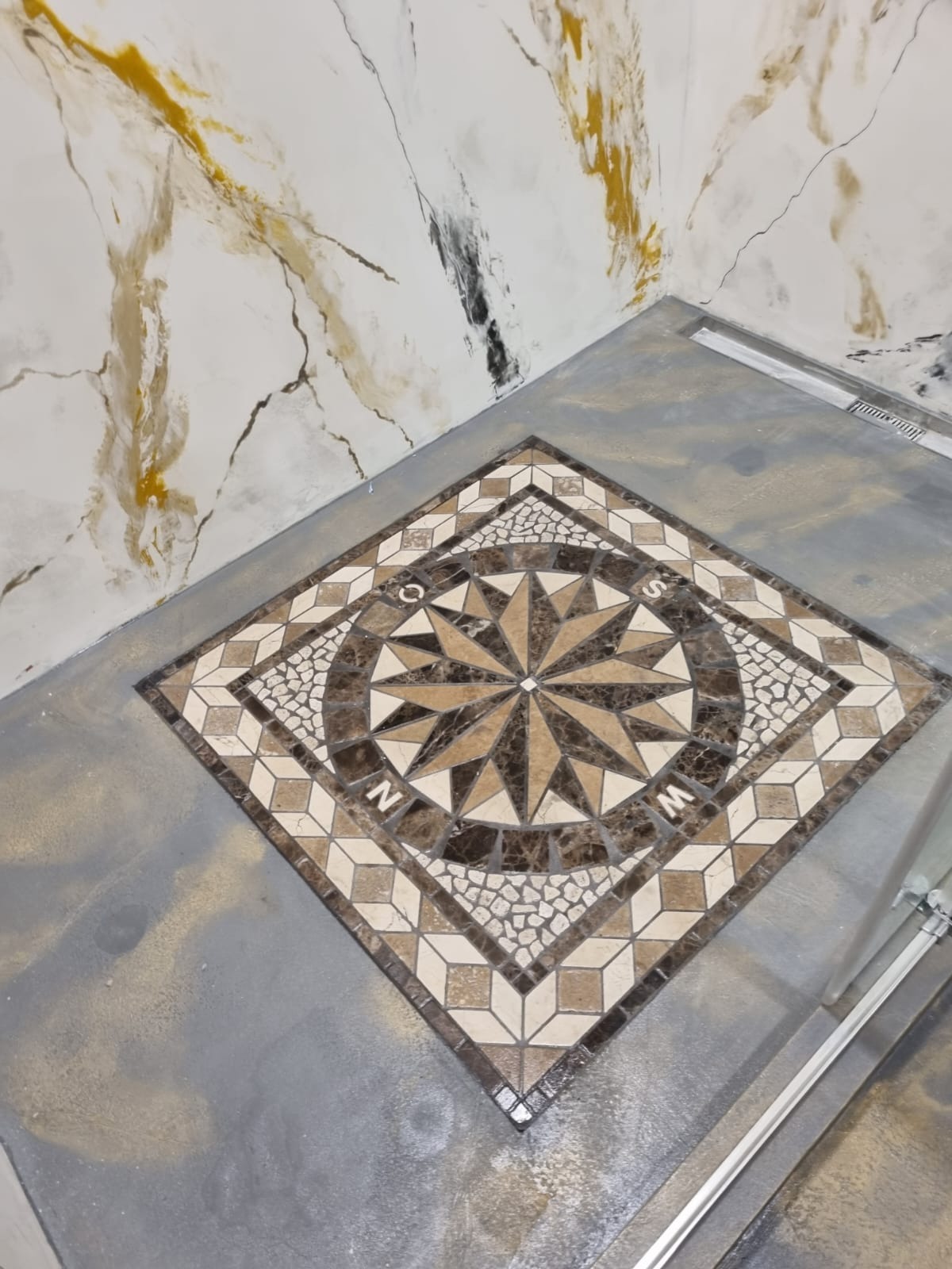 Podłoga z mozaiką w kształcie kompasu, narożnik łazienki z imitacją marmuru na ścianie i odpływem liniowym.