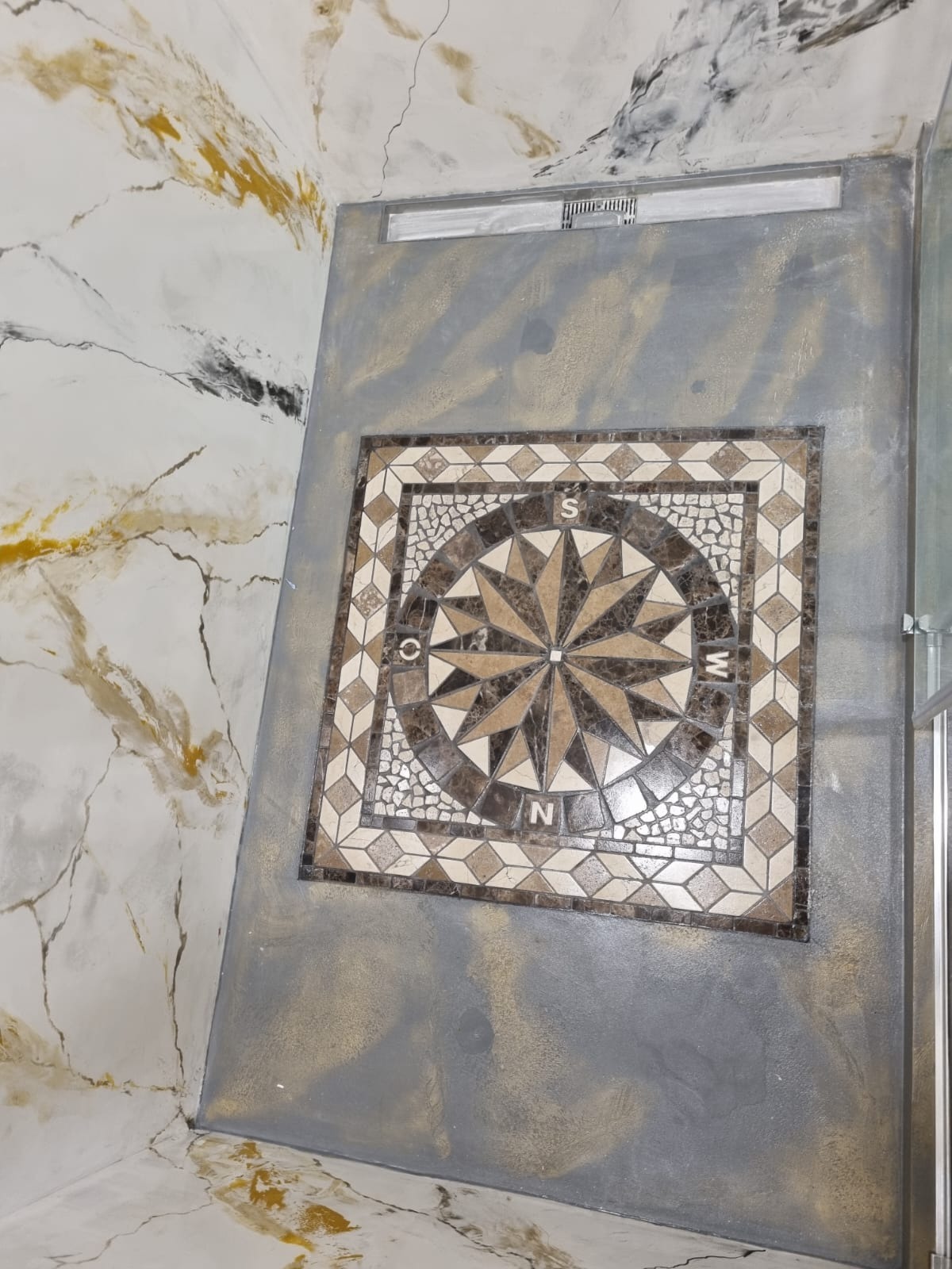 Mozaika podłogowa w łazience z motywem kompasu, otoczona szarą, dekoracyjną wylewką, ściany imitujące marmur z żyłkowaniem w odcieniach złota i czerni.
