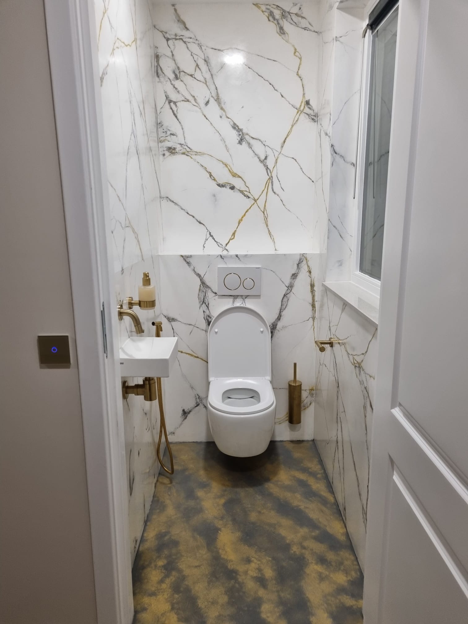Elegancka łazienka z białymi marmurowymi płytkami na ścianach, złotymi akcentami w postaci armatury i dodatków, oraz posadzką w odcieniach szarości i złota, widziana z perspektywy wejścia.