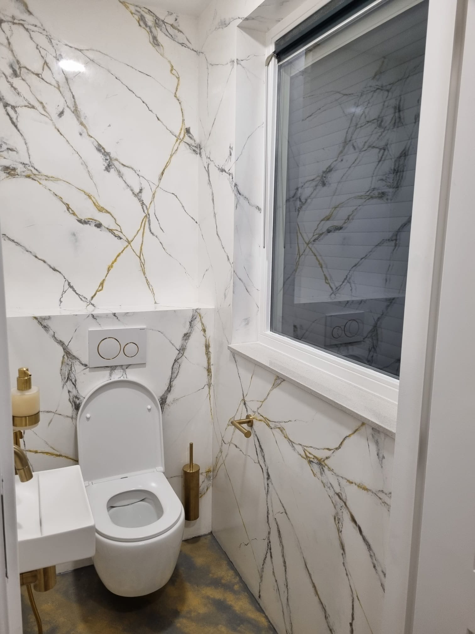 Nowoczesna toaleta z marmurowymi płytkami na ścianach, armatura w kolorze złotym, widok na okno z żaluzjami.
