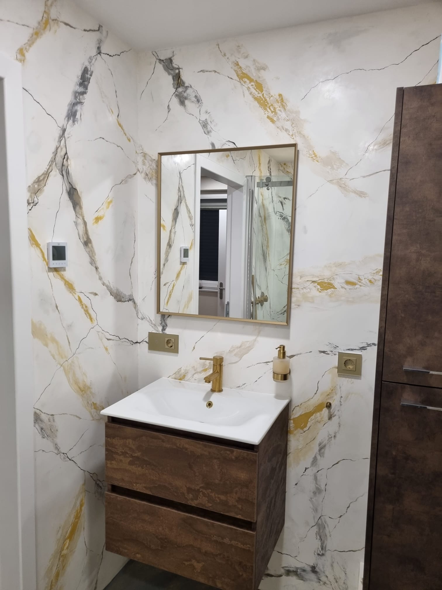Wykończona łazienka z umywalką na szafce z efektem drewna i ścianami imitującymi marmur z złotymi akcentami, widoczne gniazdka elektryczne w kolorze złotym.