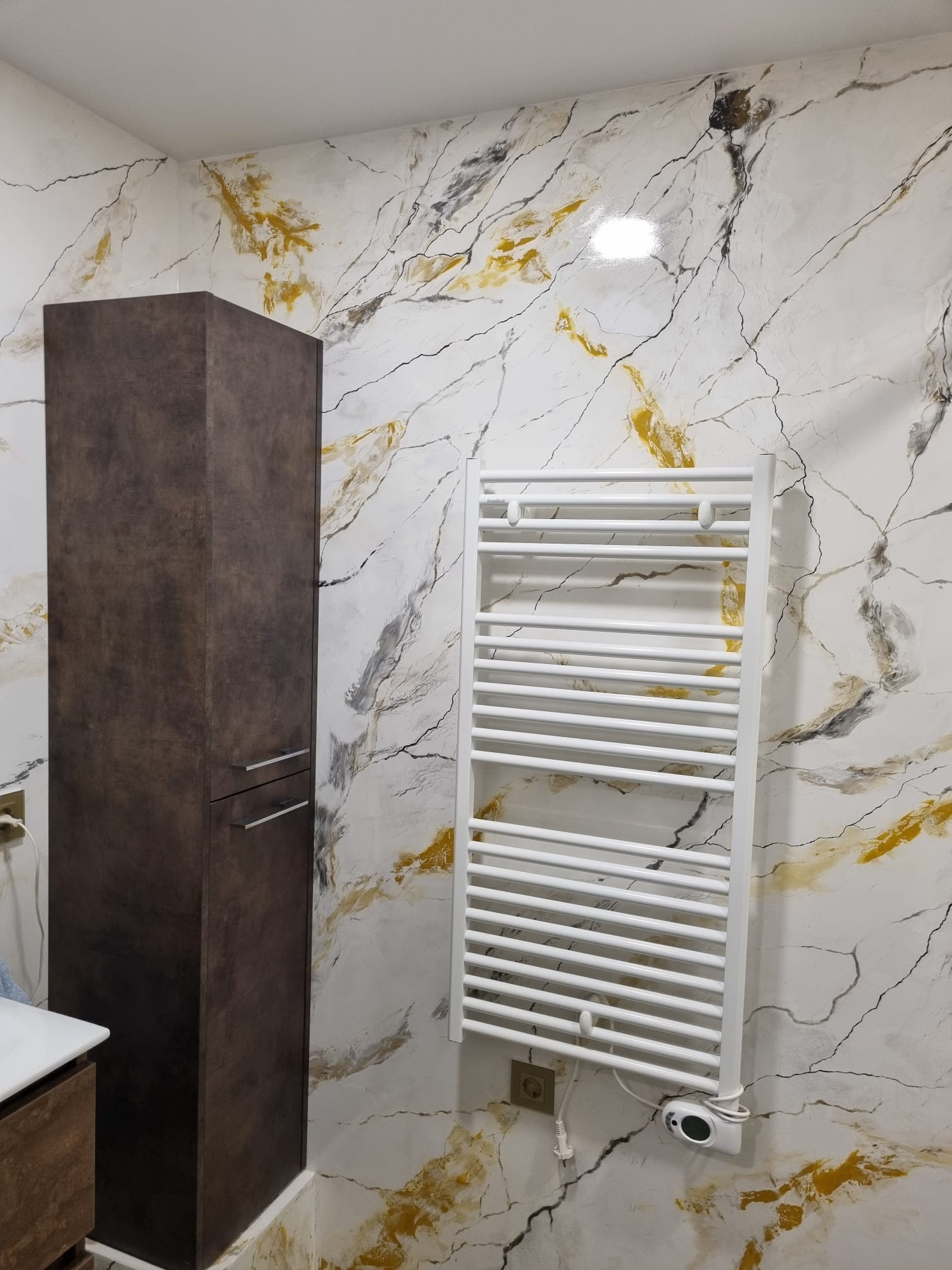 Biały grzejnik drabinkowy na ścianie z dekoracyjnym wzorem imitującym marmur z akcentami złota, obok wysoka, brązowa szafka łazienkowa z metalowymi uchwytami.