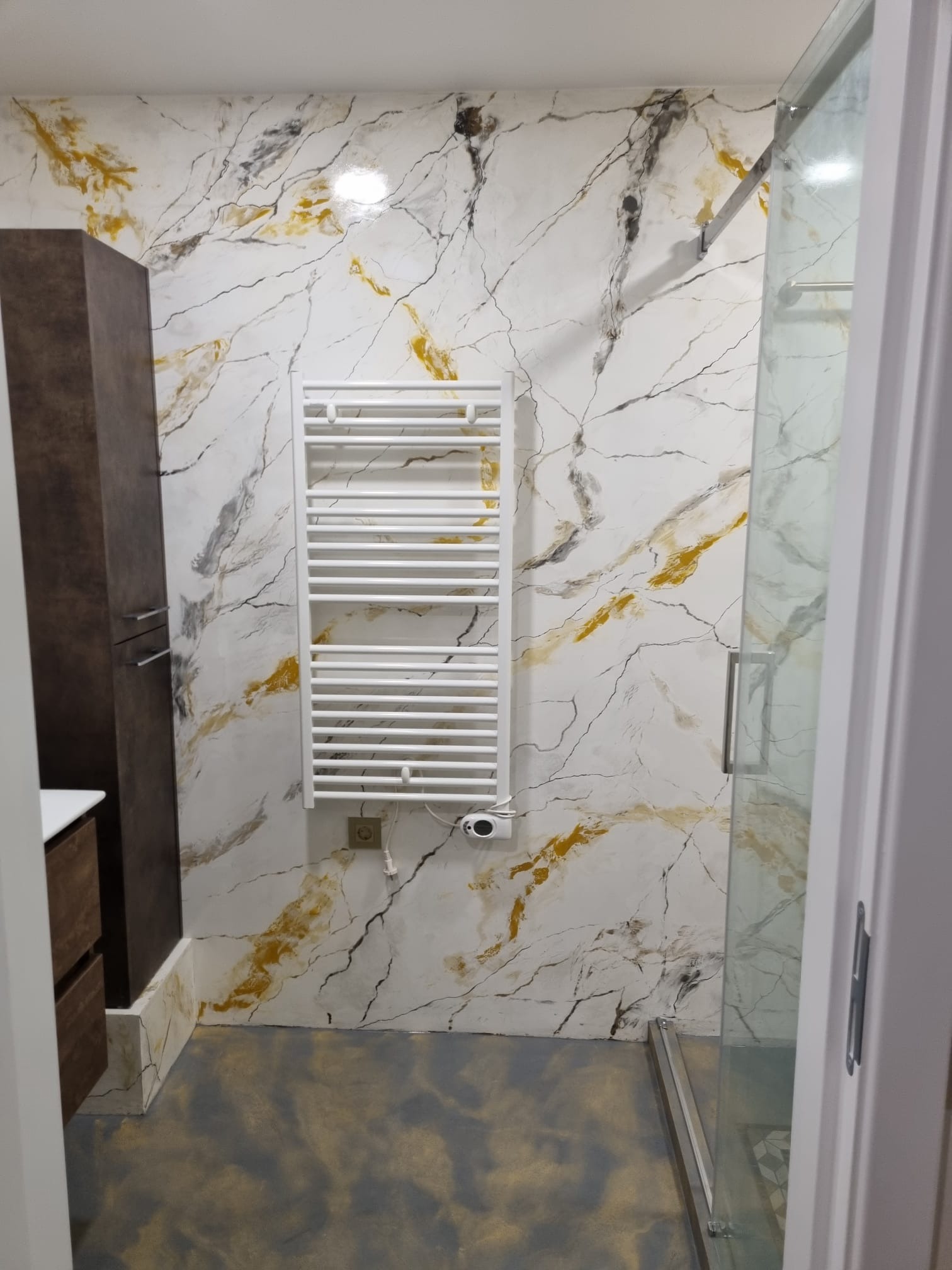 Wykończona łazienka z dekoracyjną ścianą imitującą marmur w odcieniach bieli, złota i szarości, grzejnik drabinkowy zamontowany na ścianie, ciemna szafka łazienkowa z lewej strony i kabina...