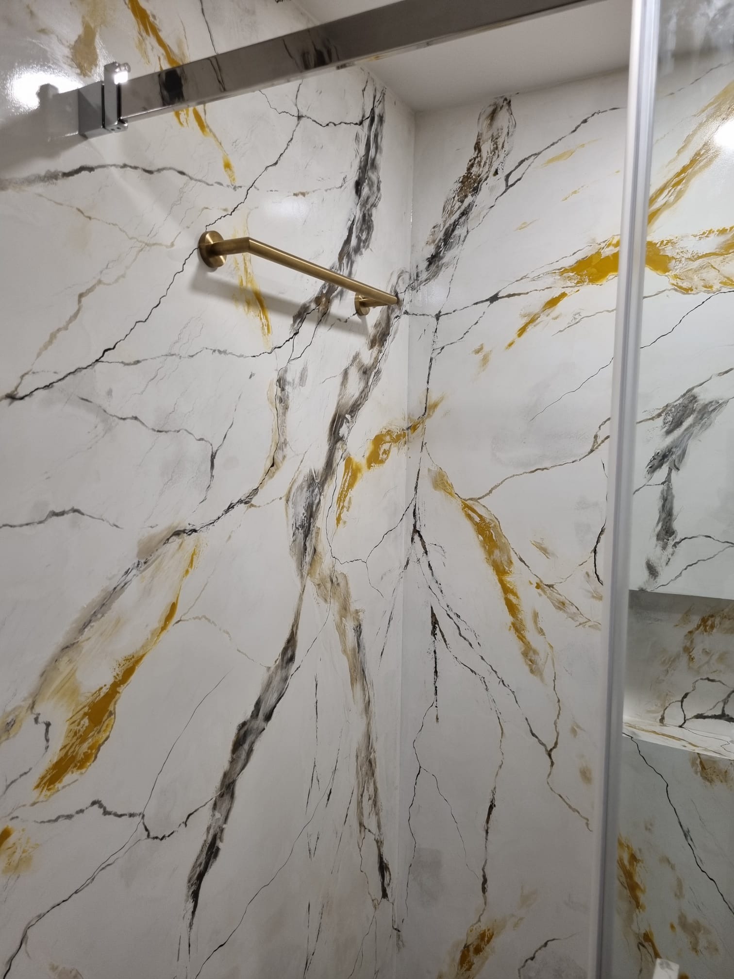 Ściana w kabinie prysznicowej z efektem marmuru w odcieniach bieli, złota i szarości, z zamontowanym złotym uchwytem na ręcznik.