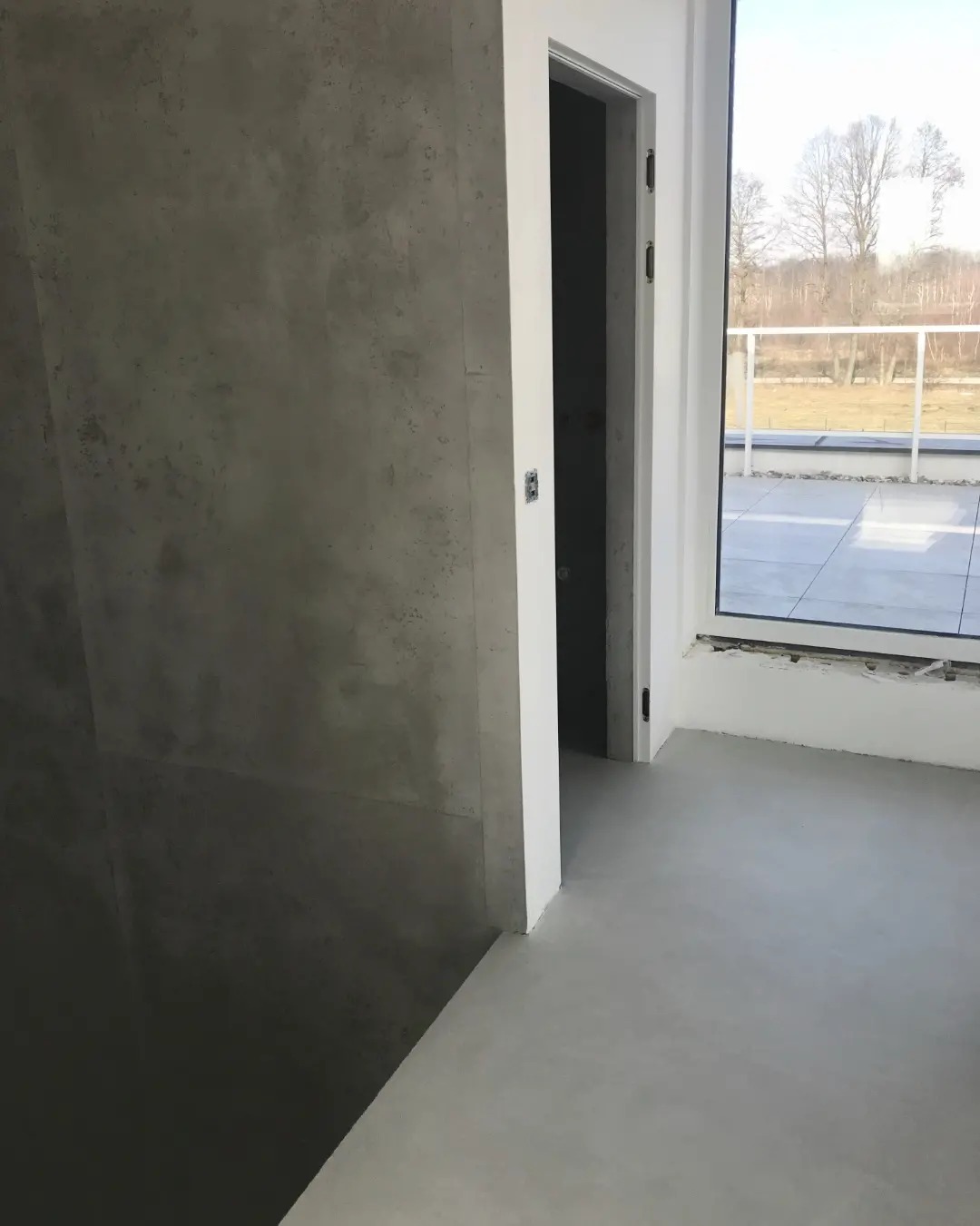 Surowe, betonowe wykończenie ściany i podłogi w jasnym wnętrzu w Warszawie, z widokiem na taras i drzewa za oknem.