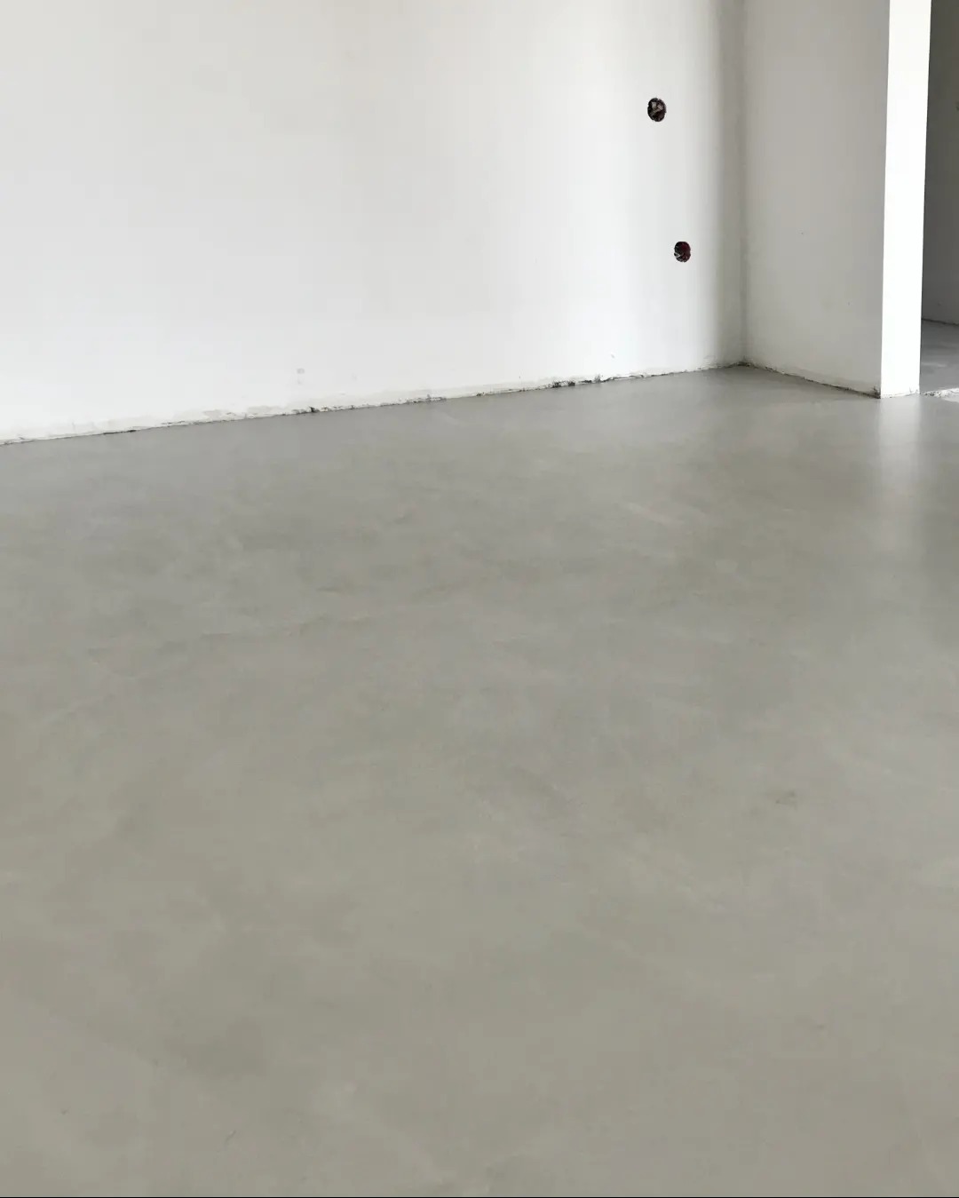 Wnętrze w stanie deweloperskim z gładką, szarą wylewką podłogową i białymi ścianami, widoczne puszki elektryczne bez osprzętu.