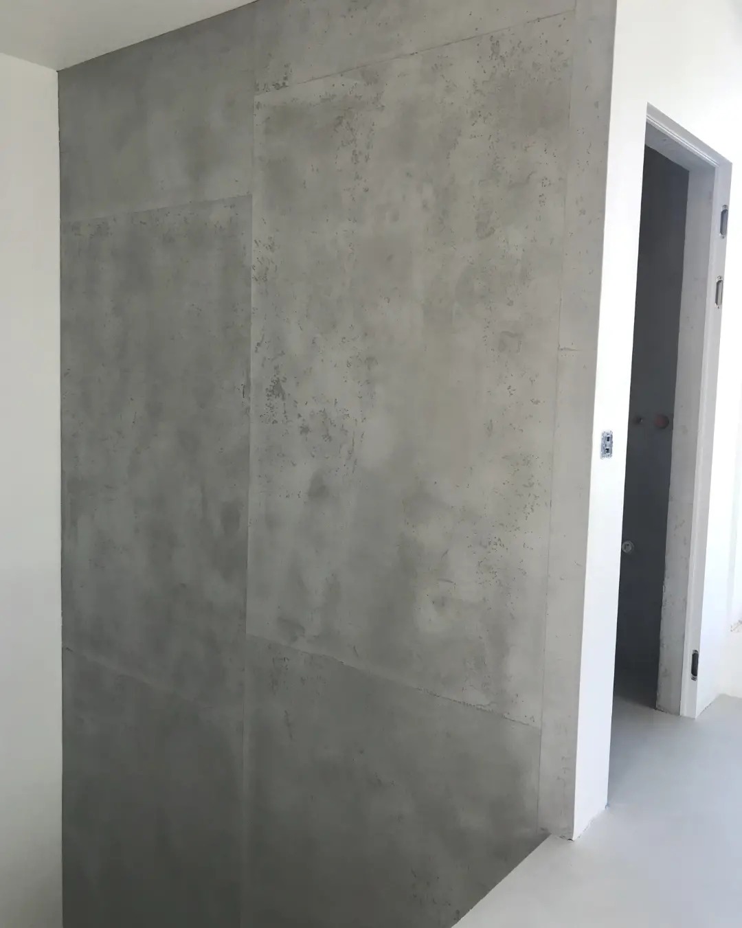 Ściana wykończona szarym, betonowym tynkiem dekoracyjnym, podzielona na segmenty, z widocznym wejściem do pomieszczenia w tle.