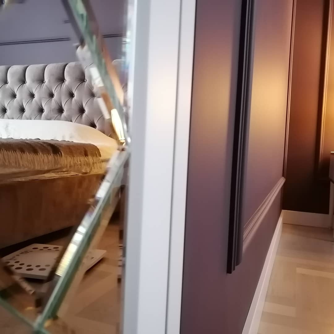 Fragment sypialni z szarym pikowanym zagłówkiem łóżka, widoczny przez lustro o geometrycznych krawędziach, oraz ciemnofioletowa ściana z białymi listwami przypodłogowymi i czarną ramą.