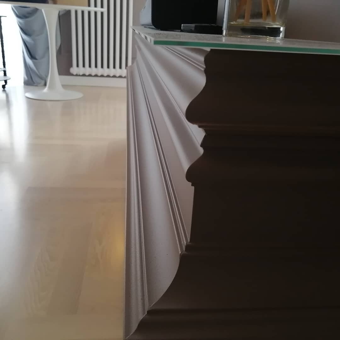 Fragment świeżo pomalowanej, ciemnej, dekoracyjnej listwy przypodłogowej z profilowanymi krawędziami na tle jasnej podłogi i białego grzejnika.