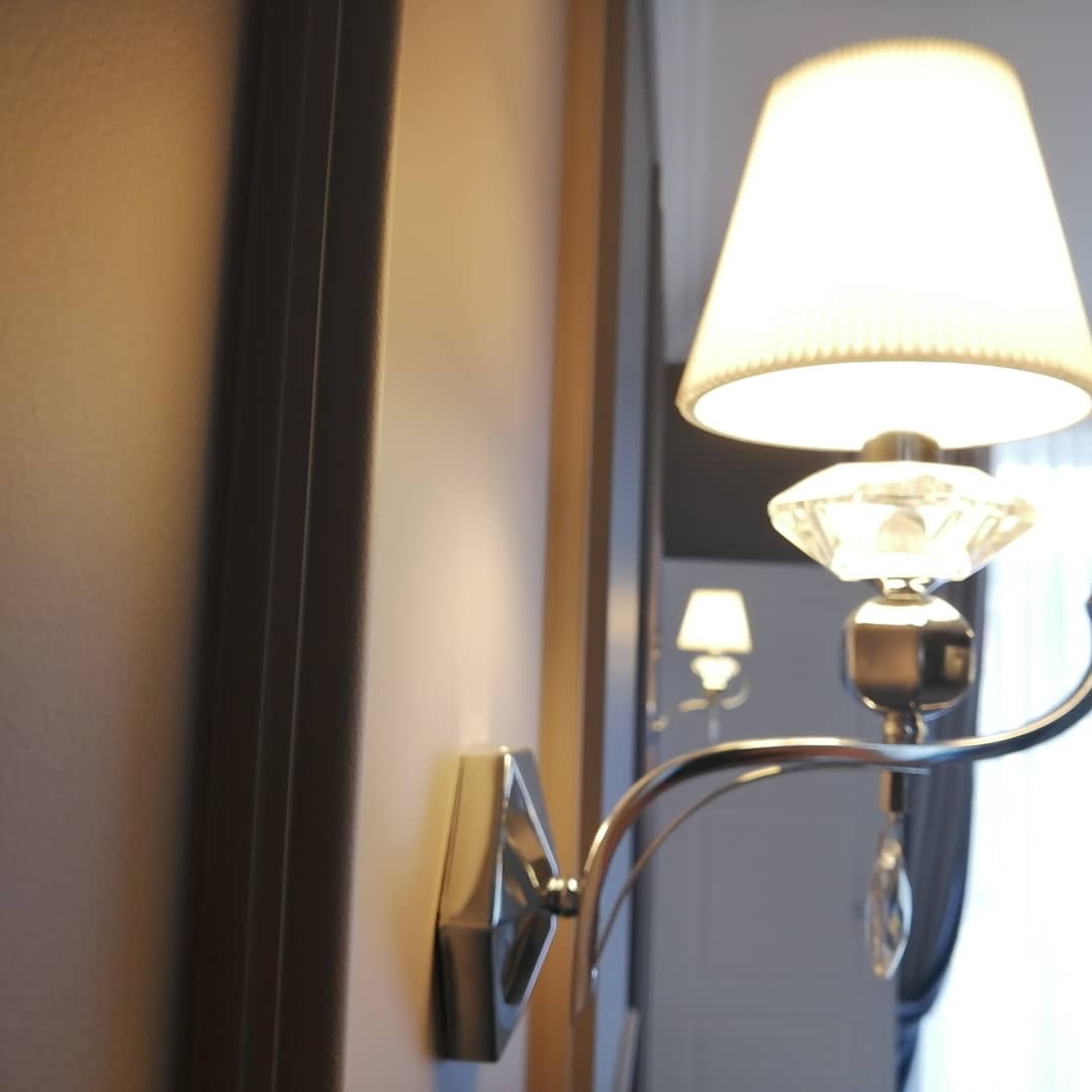 Kinkiet z kryształkami i abażurem zamontowany na świeżo pomalowanej ścianie w odcieniach beżu, widoczny fragment ramy drzwiowej.