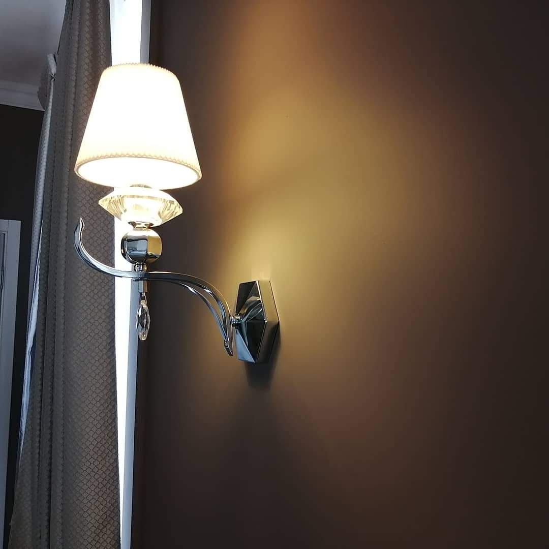 Chromowany kinkiet z kryształkami, oświetlający gładką, brązową ścianę, widoczny fragment zasłony w tle.