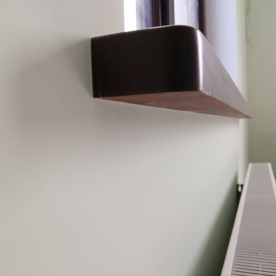 Fragment ściany z zamontowanym, ciemnobrązowym parapetem wewnętrznym o zaokrąglonych krawędziach, widoczny także fragment grzejnika.