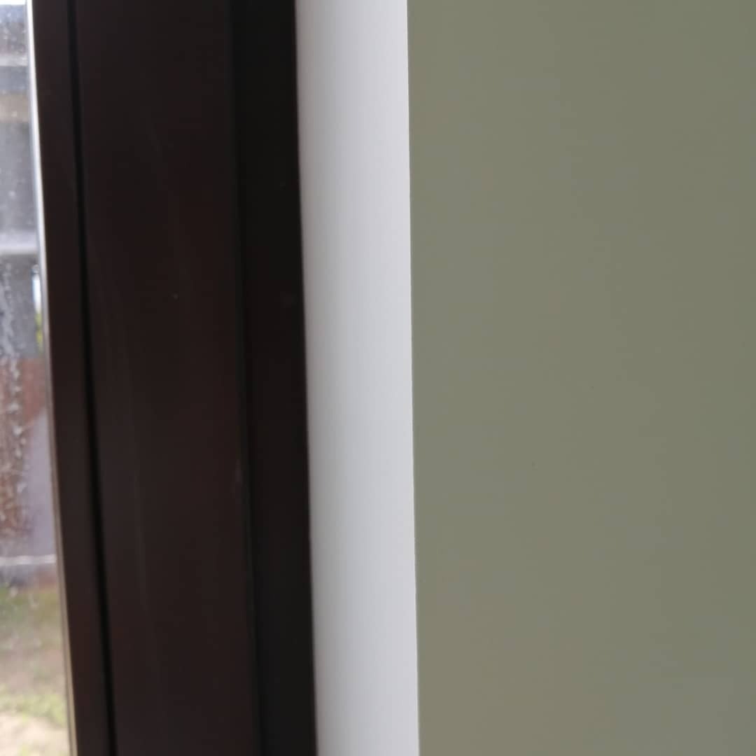 Fragment ściany pomalowanej na jasny, neutralny kolor, sąsiadującej z białym narożnikiem i ciemnobrązową ramą okienną z widocznym zamazanym widokiem na zewnątrz.