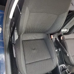 Szare, materiałowe siedzenie samochodowe z widoczną plamą na siedzisku, uchwycone z góry w słabym oświetleniu.