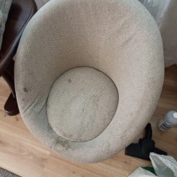 K.D - Kremowe, okrągłe krzesło tapicerowane z widocznymi plamami, obok dysza odkurzacza i butelka z płynem, ustawione na panelach podłogowych, widok z góry.