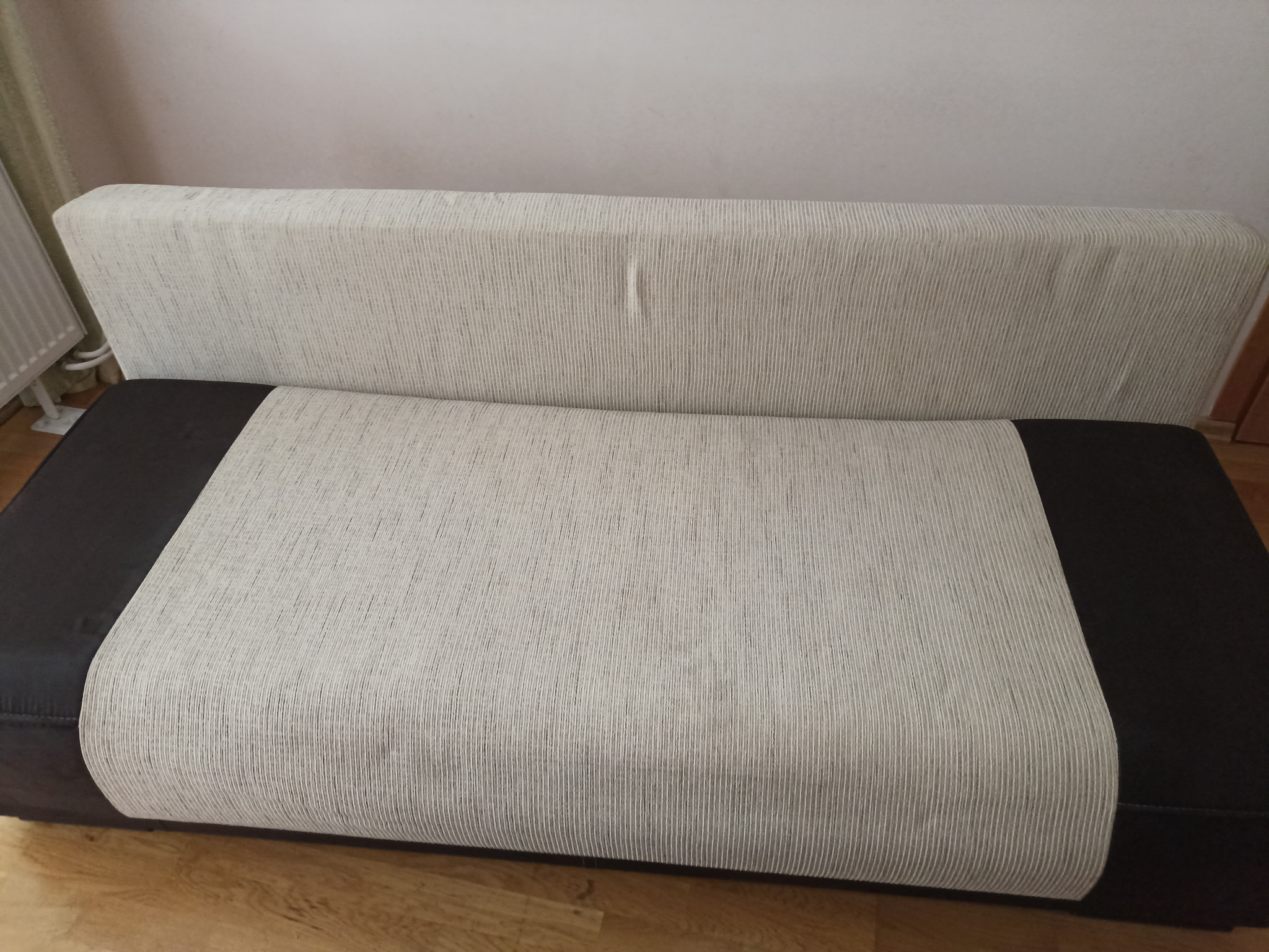 Szara kanapa z ciemnymi bokami, widoczna na tle ściany i grzejnika, z delikatnym zagniecenie na oparciu.