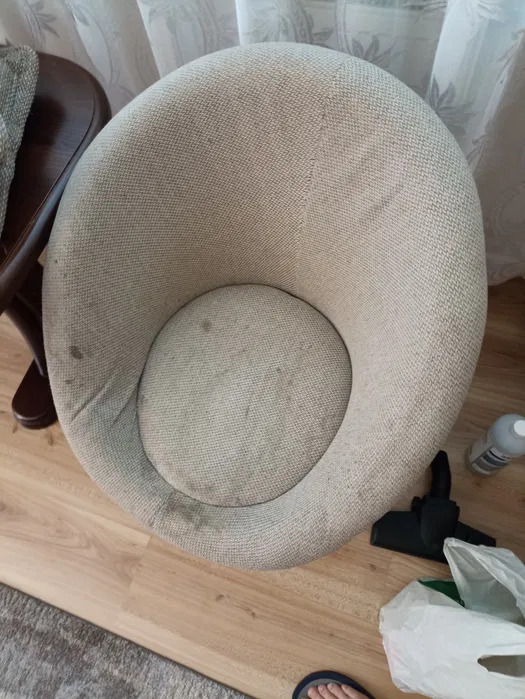 Kremowe, okrągłe krzesło tapicerowane z widocznymi plamami, obok dysza odkurzacza i butelka z płynem, ustawione na panelach podłogowych, widok z góry.