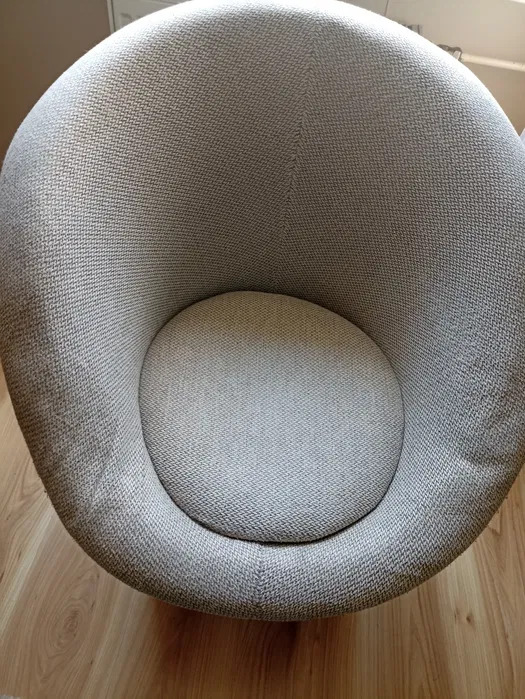 Szare, okrągłe krzesło tapicerowane z tkaniny o wyraźnym splocie, ustawione na drewnianej podłodze, widok z góry w neutralnym oświetleniu.