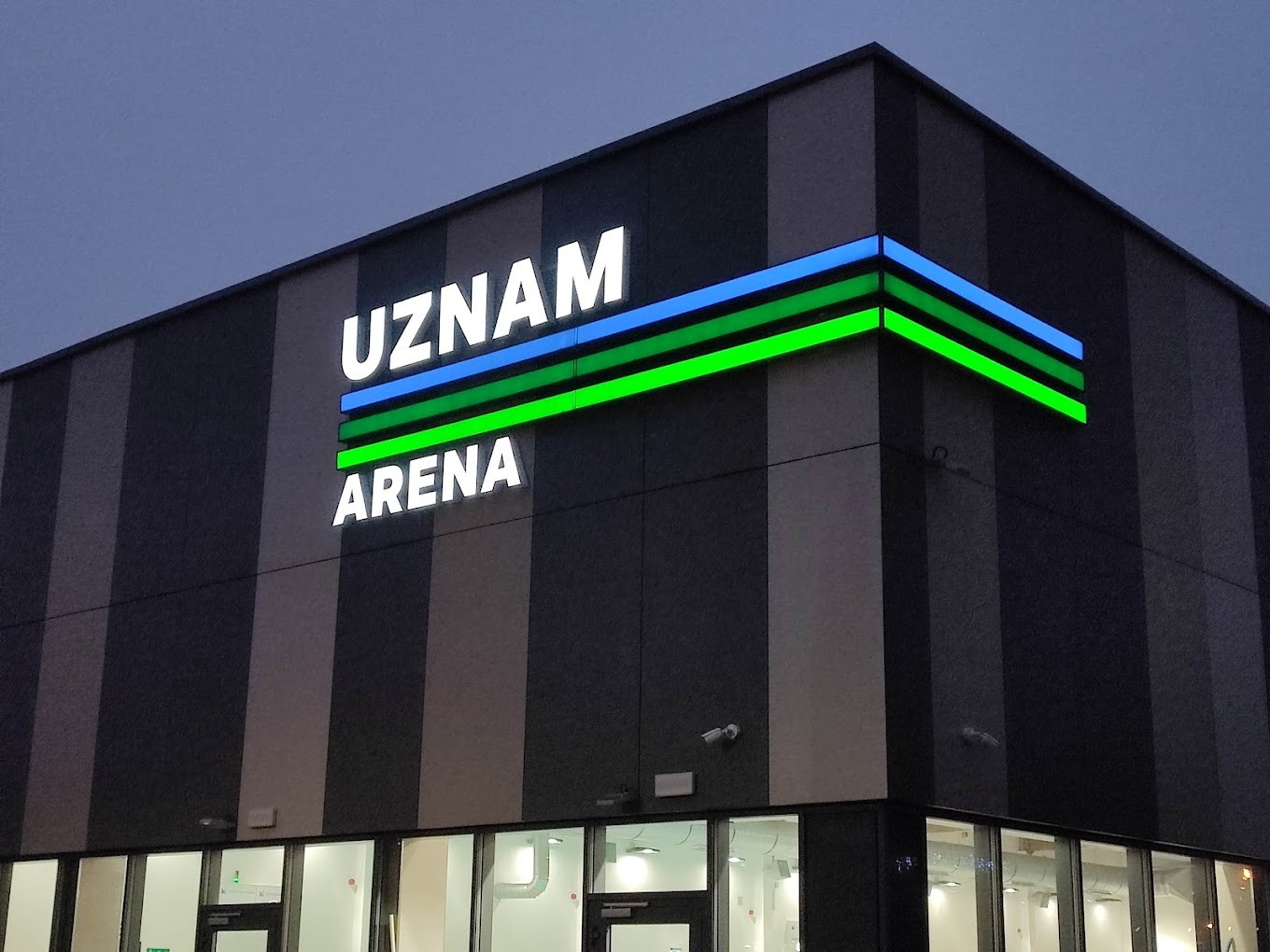 Elewacja budynku UZNAM ARENA z podświetlanym napisem i paskami w kolorach niebieskim i zielonym, widok z dołu na tle wieczornego nieba.
