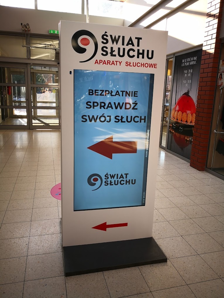 Stojący, podświetlany pylon reklamowy z logo i hasłem 'Świat Słuchu: Aparaty Słuchowe', zachęcający do bezpłatnego sprawdzenia słuchu, umieszczony wewnątrz budynku z przeszklonym wejściem.