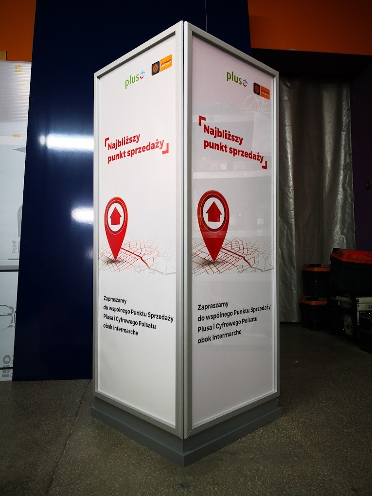 Trójstronny, wolnostojący pylon reklamowy z logo Plus i mapą wskazującą najbliższy punkt sprzedaży, umieszczony na szarym cokole.