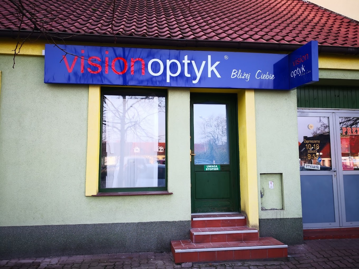 Zewnętrzny widok budynku salonu optycznego Vision Optyk z szyldem w kolorach niebieskim i czerwonym, informującym o godzinach otwarcia i hasłem 'Bliżej Ciebie'.