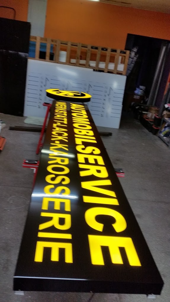 Podświetlany szyld reklamowy warsztatu samochodowego z żółtymi literami na czarnym tle, w trakcie produkcji, widoczne elementy konstrukcyjne i zaplecze warsztatowe.