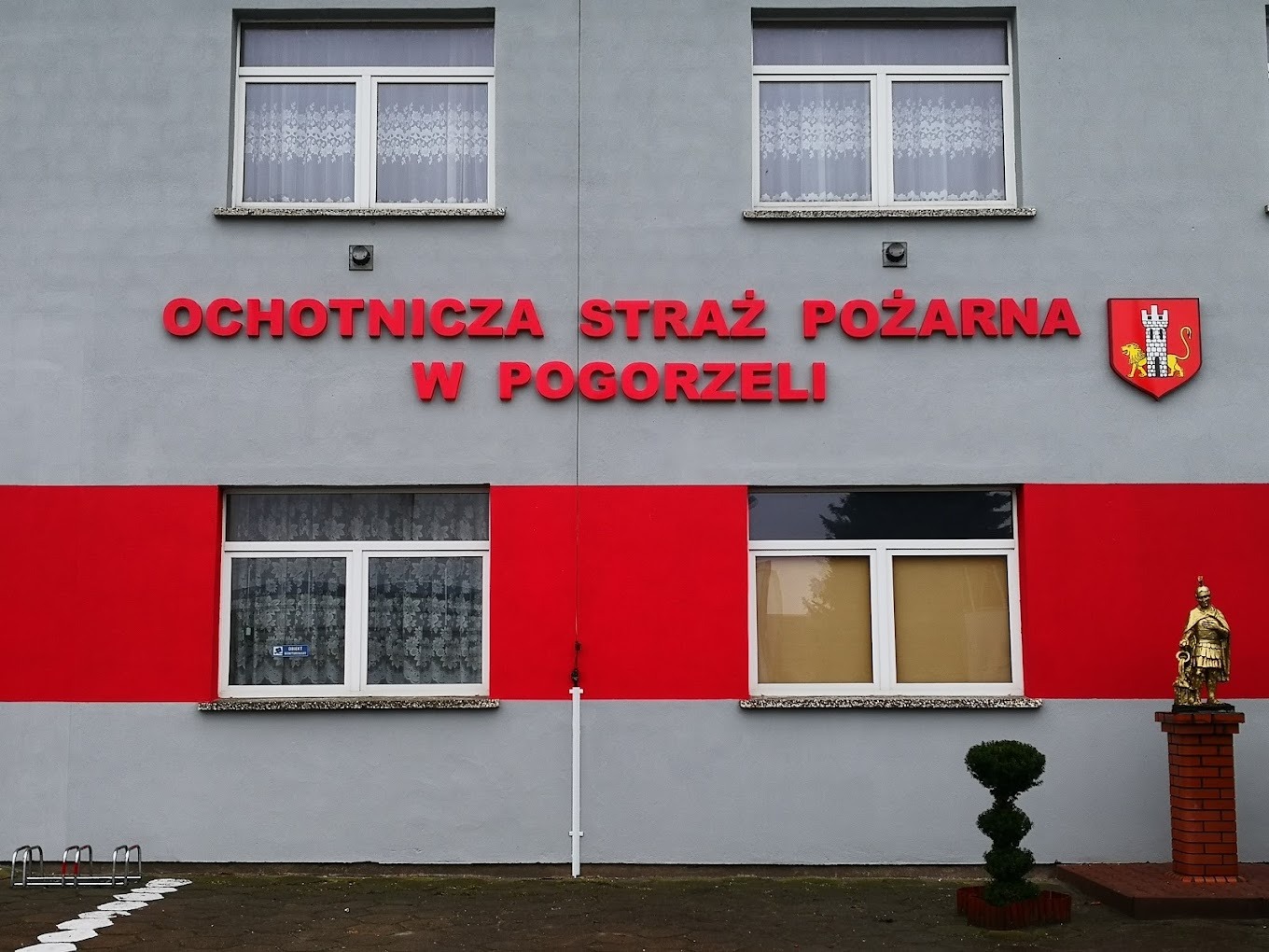 Fasada budynku Ochotniczej Straży Pożarnej w Pogorzeli z czerwonym napisem, herbem i czerwonym pasem, widoczne okna z firankami i niewielka rzeźba na postumencie.