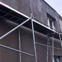 Majsterartsztukateria - Elewacja budynku w trakcie prac z użyciem rusztowania, widoczne okna i świeżo nałożony tynk w szarym odcieniu.