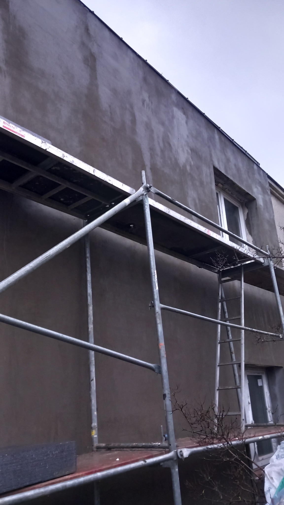 Elewacja budynku w trakcie prac z użyciem rusztowania, widoczne okna i świeżo nałożony tynk w szarym odcieniu.