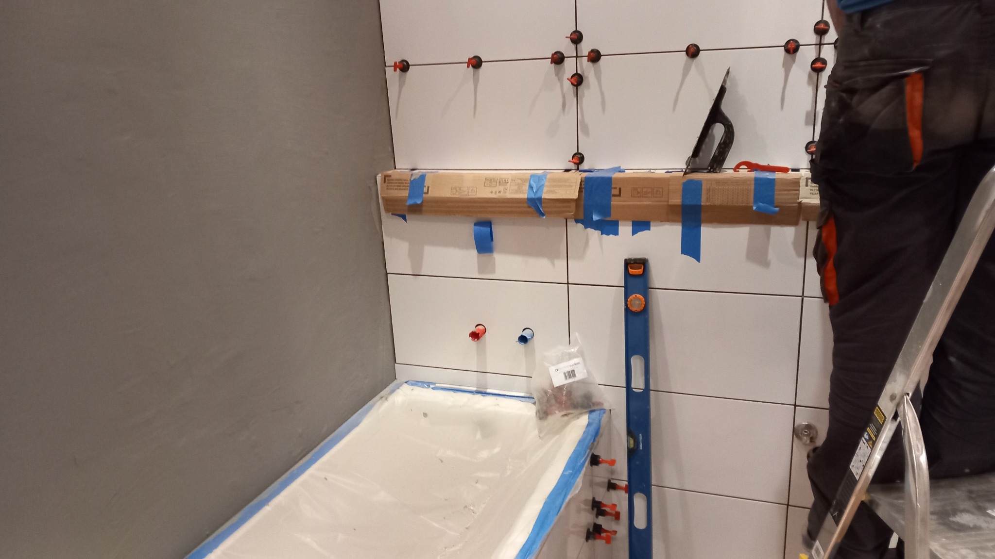 Układanie białych płytek w łazience, widoczne krzyżyki dystansowe, poziomica, wanna zabezpieczona folią i pracownik na drabinie.