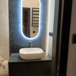 Nowoczesna łazienka z umywalką nablatową na szafce z ciemnego kamienia, podświetlane lustro owalne i szarym wykończeniem ścian.