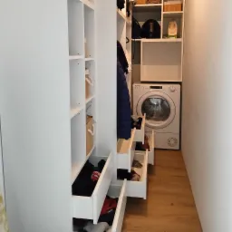 Biała zabudowa garderoby z otwartymi szufladami wypełnionymi ubraniami, obok pralka w wąskiej wnęce, drewniana podłoga.