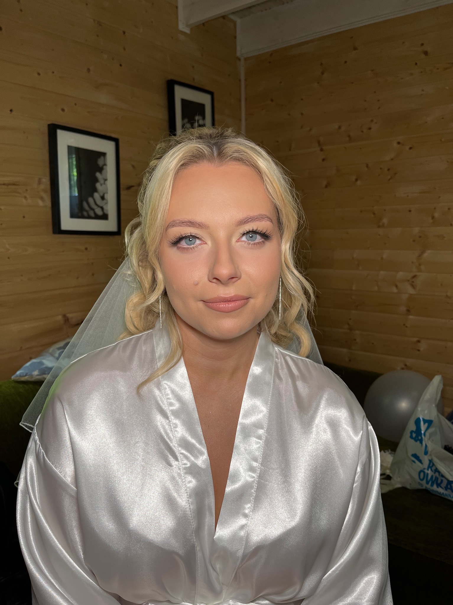 Panna młoda z blond włosami, w białym szlafroku i welonie. Delikatny, świeży look z podkreślonymi rzęsami i brwiami. Naturalne tło z drewnianą ścianą.