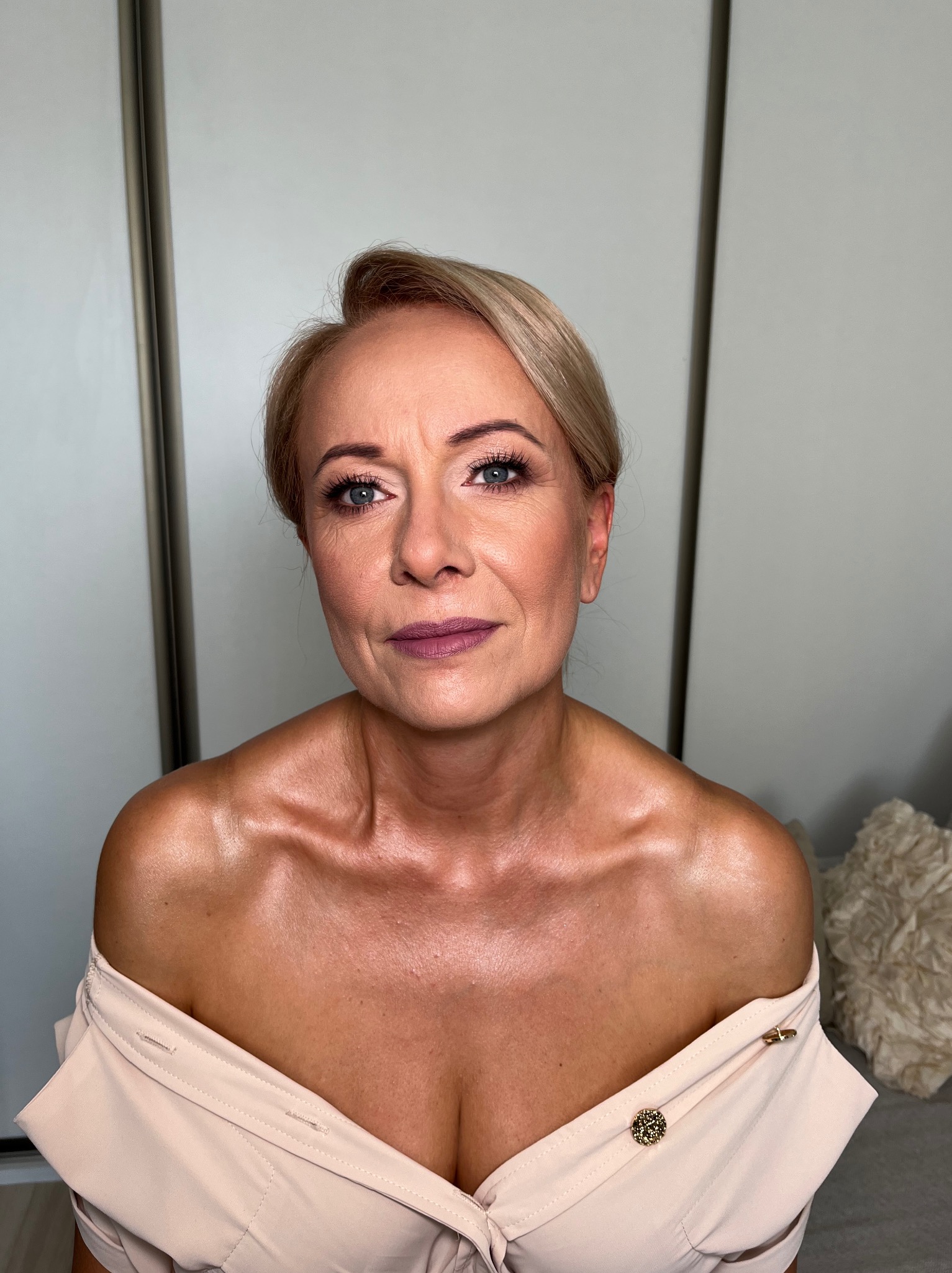 Portret kobiety z delikatnym makijażem, podkreślone oczy i usta w odcieniu fioletu. Jasne włosy upięte do góry, beżowa bluzka z ozdobnymi guzikami. Gładkie tło.