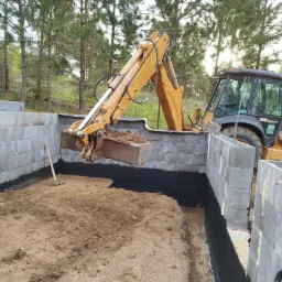 Żółta koparko-ładowarka Case 580 Super R z operatorem wybiera ziemię wewnątrz fundamentów z bloczków betonowych, zabezpieczonych czarną izolacją przeciwwilgociową, na tle drzew.