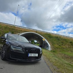 SPEED HADI TAXI MATEUSZ HADAŁA - Czarna, luksusowa limuzyna Audi A8 z tuningowanymi felgami i naklejką na przedniej szybie, zaparkowana na drodze w pobliżu tunelu z graffiti i znakiem drogowym.